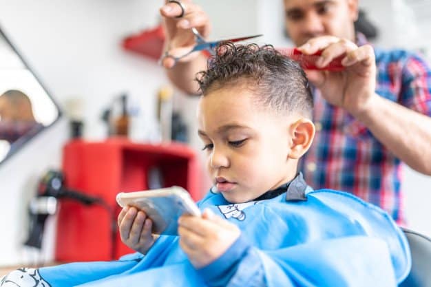 Young boy looking at a mobile phone while he is getting his hair cut