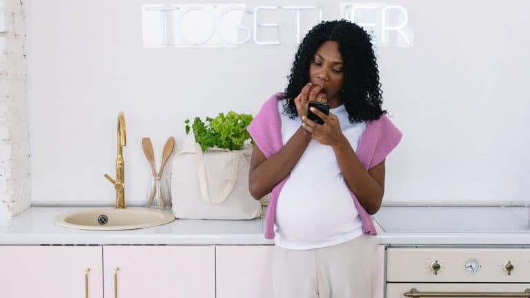Pregnant woman of colour eating apple in a kitchen while looking at smartphone