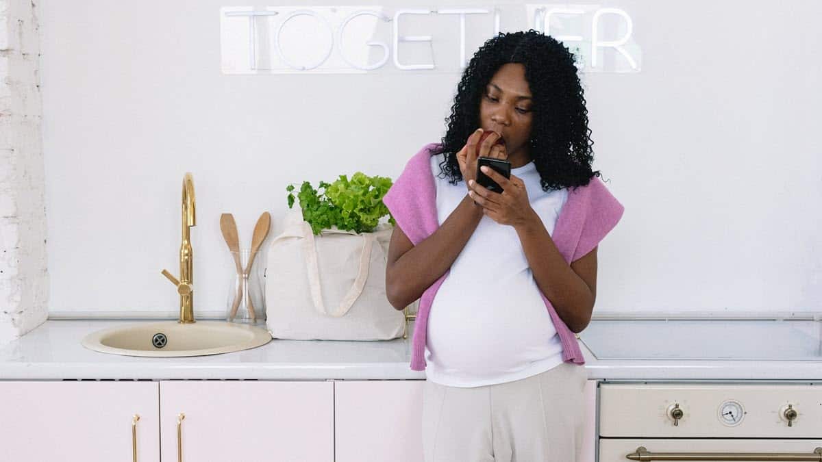 Pregnant woman of colour eating apple in a kitchen while looking at smartphone